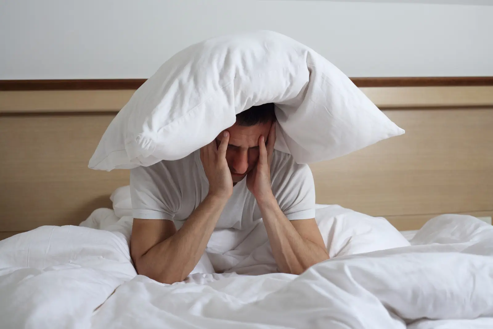 oxysphair-apnee_du_sommeil_causes Homme fatigué assis sur un lit, avec un oreiller sur la tête, souffrant de troubles du sommeil et d’insomnie.